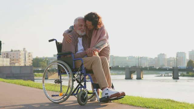 Old man in wheelchair walking with caregiver senior woman in park. Elderly family couple woman supporting taking care of paralyzed man in chair for people with disability outdoor. Rehabilitation