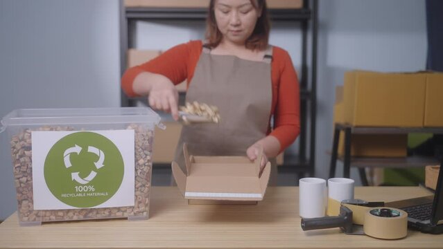 The focus is on a "100% Recyclable Materials" label, showcasing a small business's sustainable practices, A woman in the background uses eco-friendly packaging, water hyacinth cushioning
