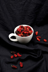 Mulberry fruit in a white cup with black fabric background. Close-up red mulberries texture. Mulberry is healthy berry fruit with a sour taste for food and drink. Fruit with black fabric background.