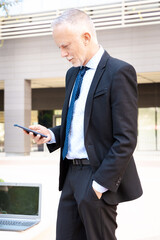 Satisfied with the results, the businessman walks down the street outside the office building, a mature boss holds a phone and coffee in his hands, writes messages and reads news online, using an app.