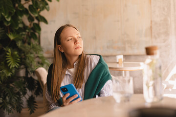 Confused puzzled woman in casual clothes, sits in cafe, holds a smartphone in her hand, looks...