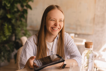 Attractive happy smiling blonde woman wear shirt sitting with tablet computer in cafe see joke and laughing. Beauty girl student using tablet computer for social media, studying or reading news.