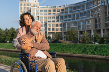 Fototapeta premium Old man in wheelchair walking with caregiver senior woman on road in park. Elderly family couple woman supporting embracing paralyzed man in chair for people with disability outdoor. Rehabilitation