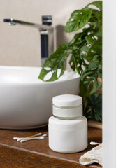 White cream jars near basin and green plant on wooden countertop in bath, mockup