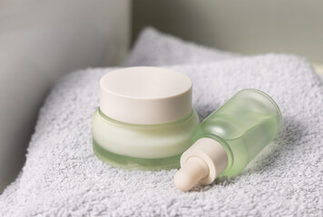 Light green dropper bottle and cream jar on light grey bath towel near basin closeup