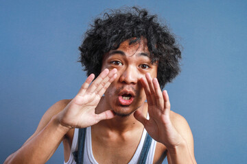 Asian Curly Man Shouting Over Blue Background. Shouting with Hands Cupped Around Mouth.