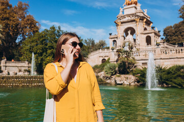 Beautiful stylish woman in trendy wear spending time on street using smartphone. Happy girl talking on the mobile phone near Ciutadella Park in Barcelona. Traveling Europe