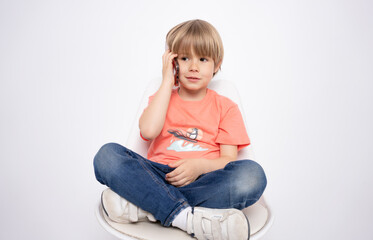 Little boy holding in hand a mobile cell phone sitting on chair isolated on white background.
