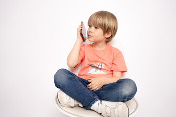 Little boy holding in hand a mobile cell phone sitting on chair isolated on white background.