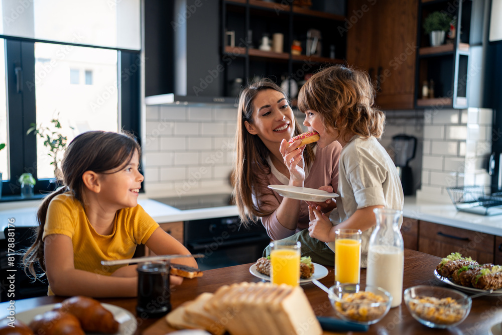 Wall mural small boy enjoying breakfast with sister and mother in the kitchen at home. happy family eating morn