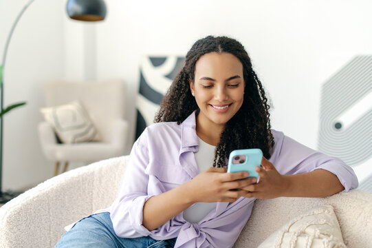 Positive attractive brazilian or hispanic stylish woman, sitting on a sofa at home in living room, uses her gadget, messaging in social media by smartphone, browsing internet, reading news, smiling