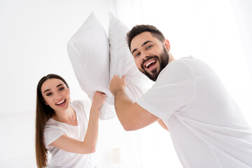 Photo of cheerful funky couple wear white t-shirts pillow fighting indoors apartment bedroom