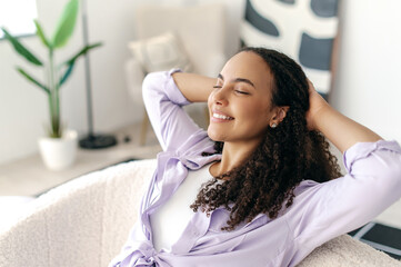 Relaxed calm curly haired stylish woman, taking a break from online work, resting with her hands behind her head and closing her eyes while sitting on a sofa, smiling