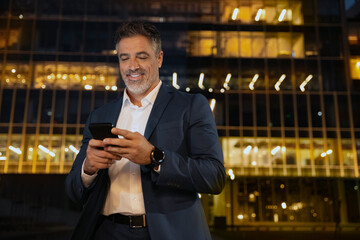 Smiling mature hispanic senior business man using app, holding smartphone cellphone at office...