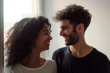 Close-up of a smiling couple looking at each other lovingly by a window, with natural light illuminating their faces.