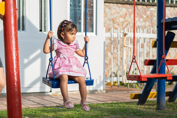 little brunette latina girl sitting on the swing in the park looking to the side while she is...