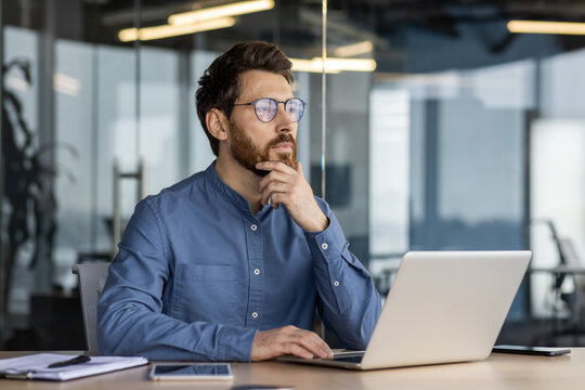 A serious and pensive young man is sitting in the office at a desk with a laptop, holding his head with his hand and looking ahead in concentration