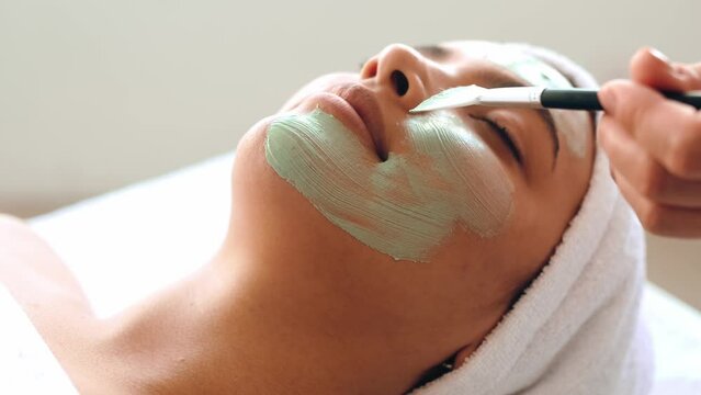Facial treatments, wrinkle prevention, facial cleansing. Therapist applying green face mask on the face of a beautiful brazilian or hispanic young woman. Skincare, beauty procedure concept