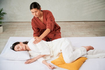 Masseuse making thai yoga massage. Woman therapist doing traditional massage treatment for female patient indoors. Client lying on mat, meditating, enjoying hand massage in calm atmosphere.
