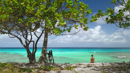 Tourist woman sitting on azure sea beach, framed by lush greenery and a tree with a bicycle leaning against, tranquility of a tropical shoreline on a clear day. Travel exotic background
