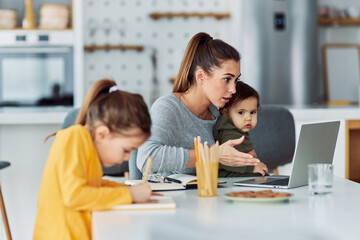 A busy working mother having a work-related video call at home on her laptop and looking after her...