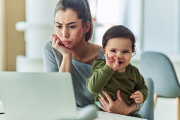 A focused mother looking over at work reports on her laptop while holding her toddler in her lap