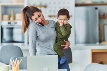 A busy mother multitasking and working from home while talking on a phone and carrying her small...