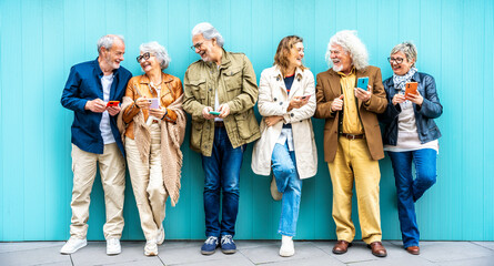 Group of senior people using technology devices together standing on a blue wall - Happy older friends having fun watching funny video on smartphone - Tech and modern elderly concept

