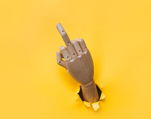 Middle finger of a right wooden hand, offensive gesture. Torn hole in yellow paper. Fuck you concept. Aggressive reaction of artificial intelligence or robot.