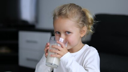 Cute girl happily drinks milk from glass at home. Child takes long sips, enjoying taste drink. Milk is child favorite drink and source of important nutrients. Happy face radiates joy from healthy milk