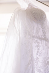 A close-up of a white lace wedding dress hung up by a window. 