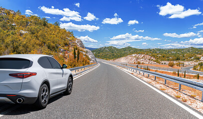 A white car drives along the highway against the backdrop of rocky mountains on a sunny day.