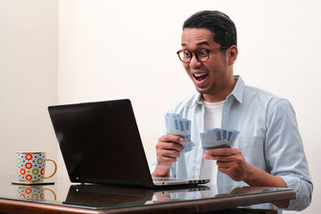 A man looking to his laptop with amazed expression while counting money