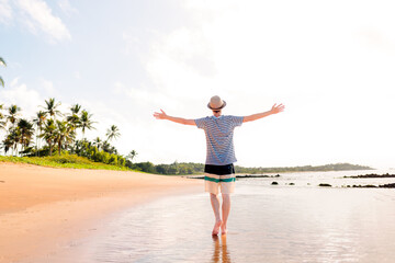 Brazilian man wearing sun hat, with his arms raised to express gratitude and happiness for enjoying summer vacation on a paradise beach
