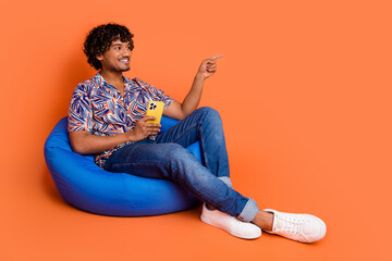 Full length photo of handsome man sit beanbag point empty space device dressed stylish colorful garment isolated on orange color background