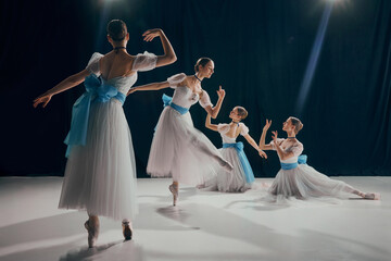 Four young dancers in ethereal white tutus and blue sashes, posing elegantly on dimly lit stage. Choreography. Concept of beauty, classic and modernity, contemporary art. Ad