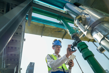 Male engineer monitoring machinery on a rooftop with city skyline in the background, wearing safety...