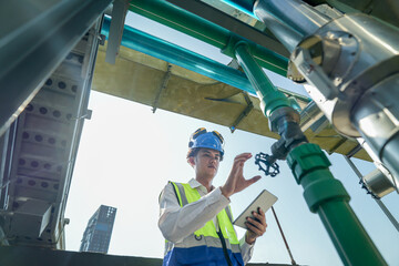 Male engineer monitoring machinery on a rooftop with city skyline in the background, wearing safety...