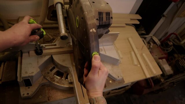 POV shot of unrecognizable handyman with arm tattoos working with miter saw on wood on workbench in carpentry workshop