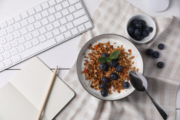 Delicious granola with blueberries in bowl, stationery and computer keyboard on white wooden table,...