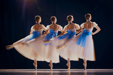 Rear view of young girls, ballet dancers in white tutus with blue bows on pointe dancing in motion...