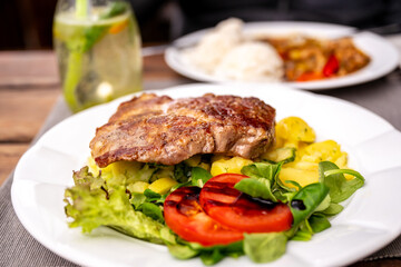 Pork steak on potato salad with vegetable on first white plate, drink and second meal on table.