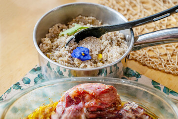 Buckwheat with flower in pot, spoon, part of baked veal on table.