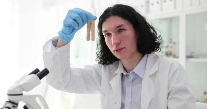 Male employee with curly hair looks at samples in tubes sitting at table in modern office. Chemist compares elements doing experiment slow motion