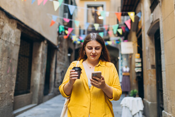 Happy cheerful young woman with paper coffee cup walking on city street checks her smartphone....