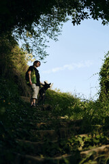Young woman with black mix breed dog on an old stone staircase in the middle of a green spring park. A dog with female owner rests outside on a summer sunny day in shadows of trees.
