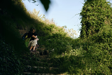 Young woman with black mix breed dog sits and poses on an old stone staircase in the middle of a green spring park. A dog with female owner rests outside on a summer sunny day in shadows of trees.