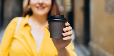Woman gives paper black coffee cup or tea on urban background. Take away or delivery concept. Copy...