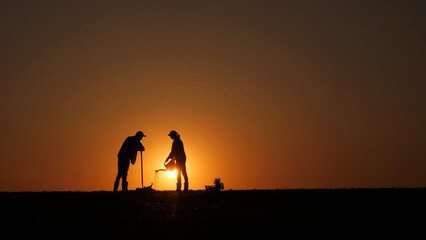 A couple of farmers plant seedlings in a field at sunset. A man digs, a woman waters