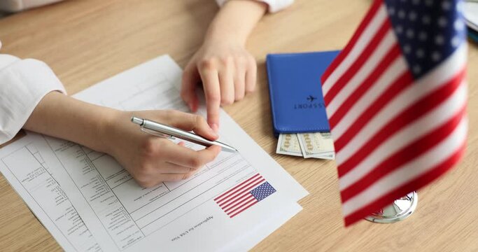 Female person fills in visa application form in room. Woman signs documents sitting at wooden desk with USA flag and passport with dollar bills slow motion